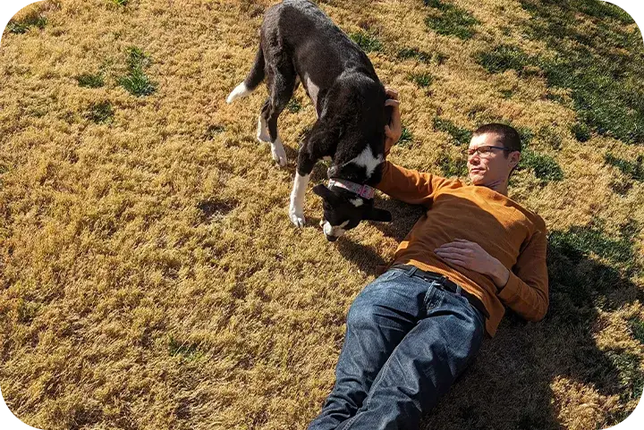 TJ Robertson lying on the grass in an orange sweater, laughing, with his dog standing over him.